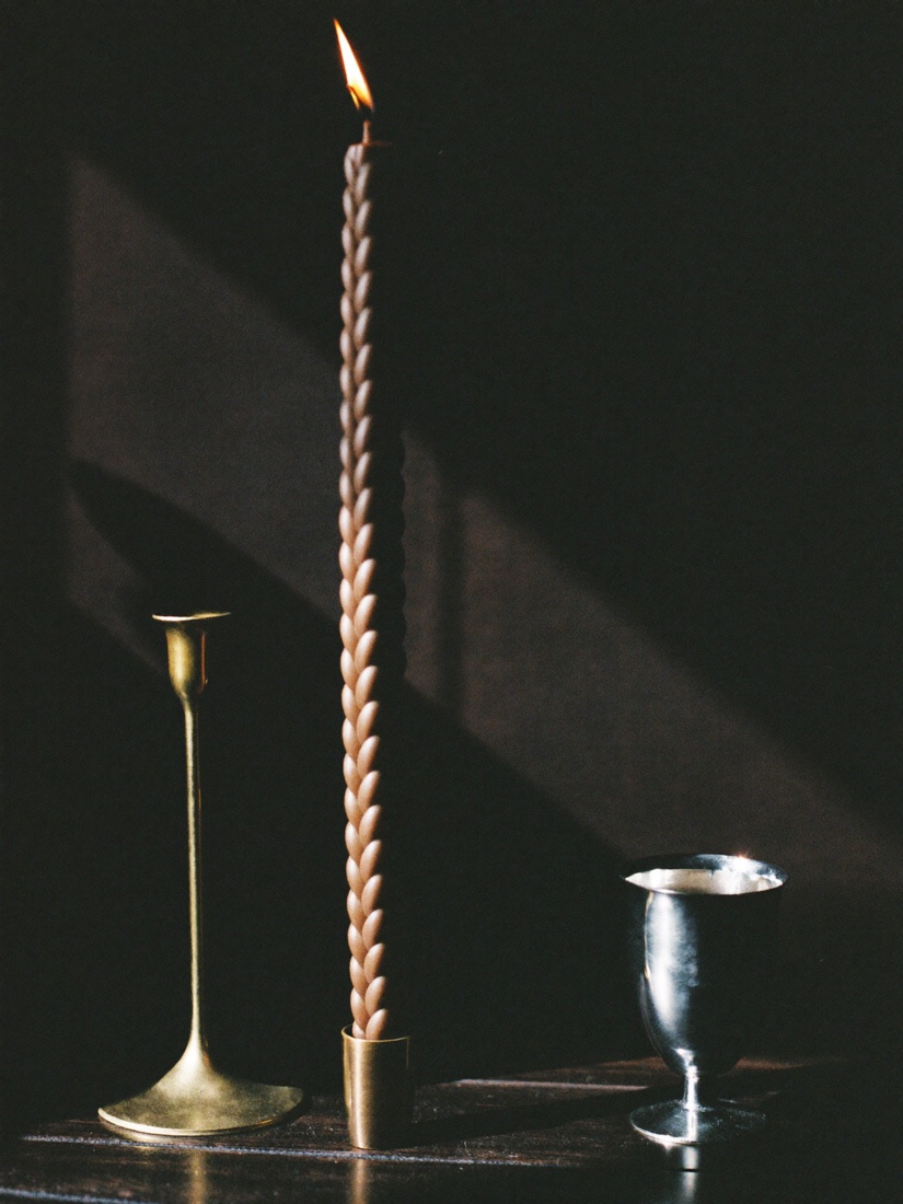 A tall braided havdalah candle burning beside a brass candlestick and a silver wine cup